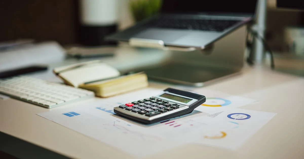 Calculadora sobre planilhas em uma mesa de escritório, com um notebook ao fundo