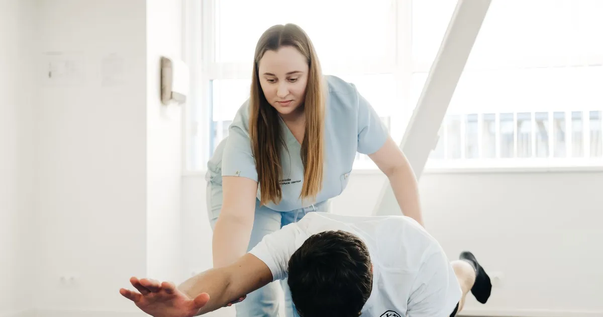 Fisioterapeuta de uniforme azul-claro ajudando um homem deitado de bruços a esticar o braço e a perna em uma sala clara